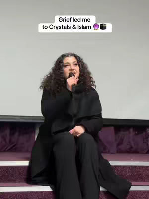 A woman with curly brown hair is seated on a purple cushioned surface, holding a microphone. She is wearing a black coat and pants. Above her is a text overlay that says, 'Grief led me to Crystals & Islam'.