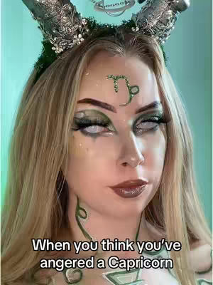 A woman with long blonde hair, wearing elaborate Capricorn-themed makeup. She has white contact lenses, dark eye makeup, and a green Capricorn symbol on her forehead. She wears a headdress with ornamental horns and green leafy accents and has zodiac signs drawn on her neck.