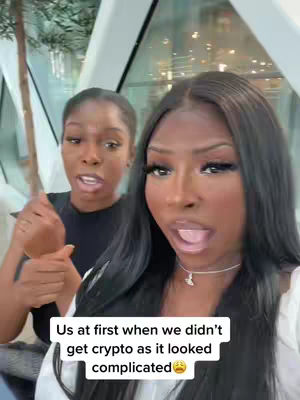 Two women are positioned in front of a bright, modern backdrop. The woman on the right, with long, dark hair, has her mouth open while looking toward the camera. The woman on the left is partially visible, also with her mouth open. Overlay text at the bottom reads, 'Us at first when we didn't get crypto as it looked complicated'.