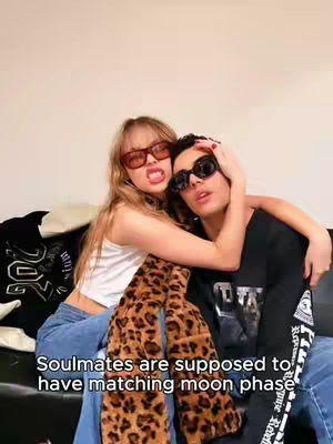 A young woman with blonde hair and red-tinted sunglasses is seated next to a person with dark hair and black sunglasses on a black sofa. The woman is smiling with her mouth open. The man has his arm around the woman. The woman is wearing a white tank top, blue jeans and furry, leopard-print boots. The man is wearing a black t-shirt and blue jeans. Text at the bottom says, "Soulmates are supposed to have matching moon phases."