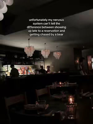 The image captures a dimly lit restaurant scene, focusing on the interior. Gentle light emanates from several large, ornate chandeliers suspended from the ceiling. Below the lights, tables are set with glassware and place settings. The back of the restaurant features a bar area with bartenders visible. Text overlays the image.