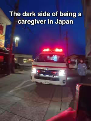 A nighttime scene featuring an ambulance with flashing red lights parked on a street. In the foreground, a hand holding a phone is visible, implying a video recording. The text overlayed reads "The dark side of being a caregiver in Japan". The composition suggests a real-life event or situation related to caregiving.