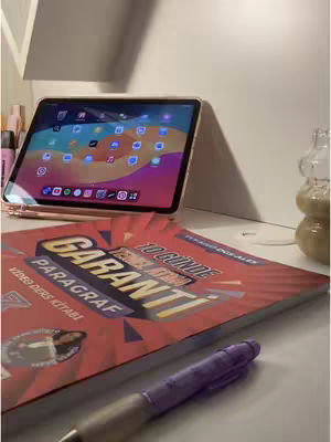The image shows a study setup with a laptop, a visible textbook, and a pen. The laptop is angled to display a bright screen, and the book is open displaying red and orange colors. A pen lays on a white surface near the red book and an iPad in the back. The layout suggests a study-related setting.