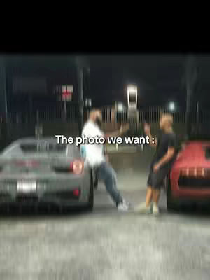 The cover image captures a nocturnal scene with two men flanking sports cars. Text overlayed reads 'The photo we want'. Two men, one in a white shirt and jeans gesturing with an outstretched arm, and the other in a black shirt, stand by the cars. The cars are positioned with one being grey and another being red. The background is blurred, implying a busy environment with visible lights.