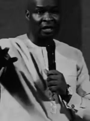 A grayscale shot of a man, Apostle Joshua Selman, speaking into a microphone. He's wearing a light-colored shirt and a watch. The background is blurred, focusing attention on the speaker and the microphone, suggesting either a sermon or a motivational speech.