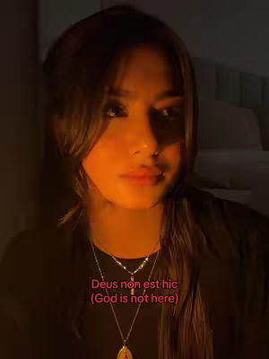 A medium shot features a young woman with brown hair and a contemplative expression. Her face is softly lit, with the lighting coming from the bottom, casting shadows. She wears two necklaces and a black top. Text overlayed in pink states, 'Deus non est hic (God is not here)'.