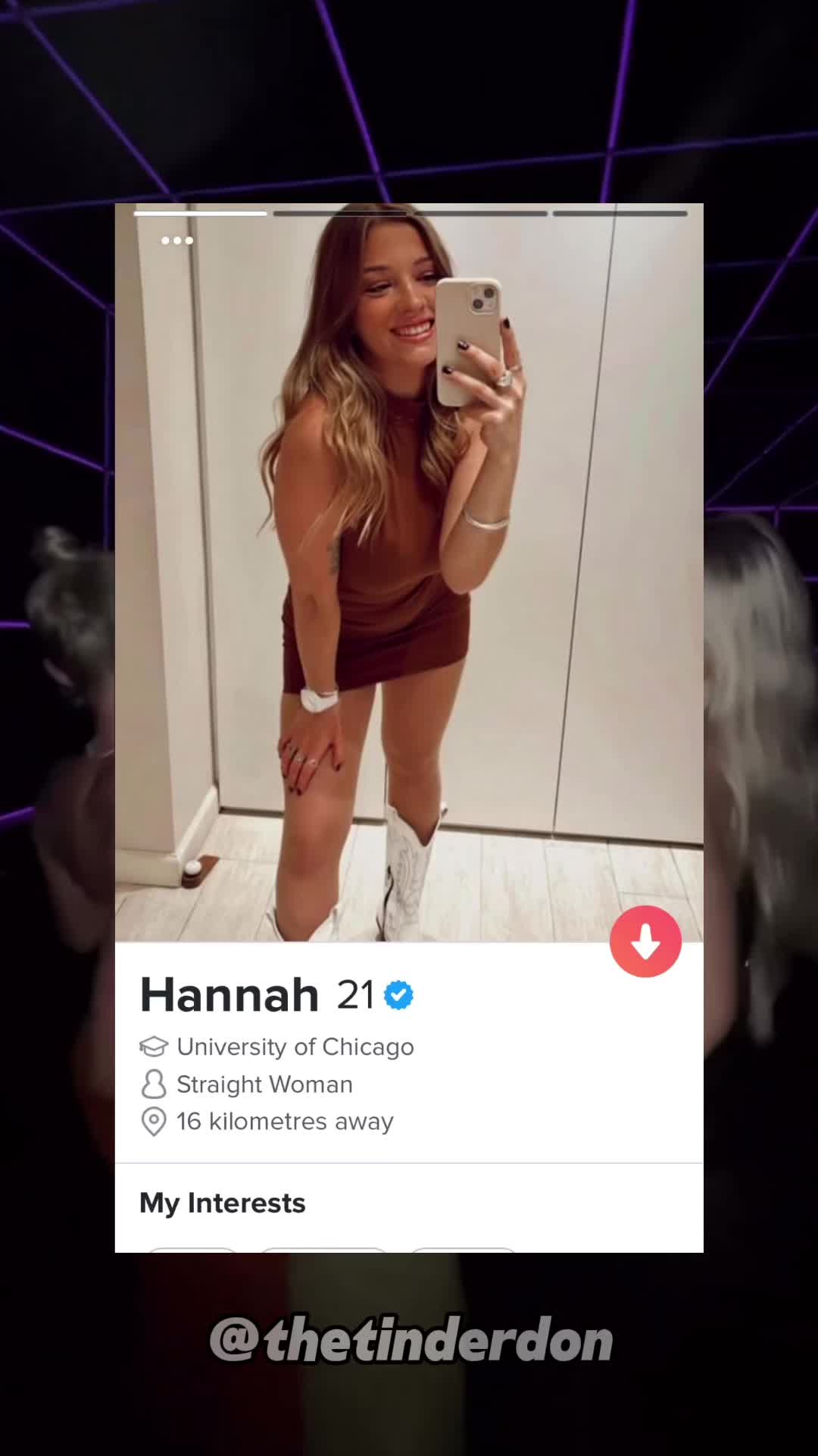 A young woman is shown holding her phone up in a mirror, taking a selfie. She is smiling and wearing a brown dress and white boots. The image is framed within a Tinder profile interface, with a purple background suggesting the video has been edited.