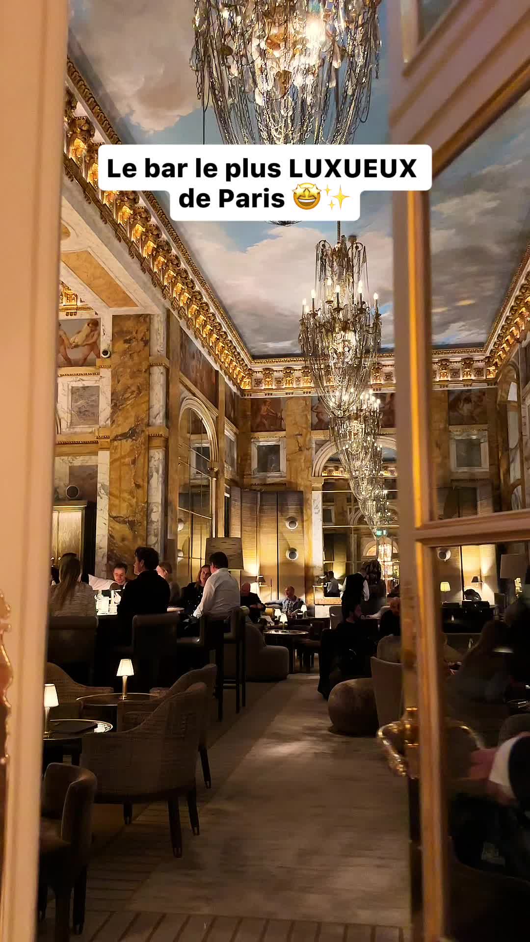 The cover image showcases the interior of a luxurious bar, possibly in a hotel. The ceiling is painted or decorated with a blue sky, and grand chandeliers hang down, illuminating the scene. The bar is filled with patrons seated at tables and the bar, giving the impression of a lively and upscale atmosphere.