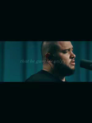 The cover image shows a vertical composition. The foreground features a man with a shaved head and a beard facing slightly right, singing into a microphone. He's wearing a dark top, and an earpiece is visible. Faint text is overlaid, on his left, likely a quote related to the song. The background is blurred, suggesting a studio or performance setting, and colored in cool shades of blues and greens. The overall lighting is focused on the man's face, making the composition dramatic and intimate. The color contrast is very slight.