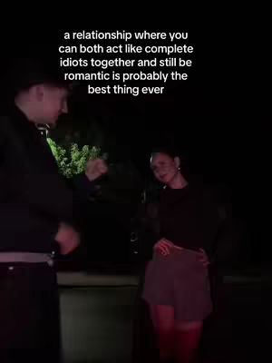 A nighttime shot with two figures in the foreground, one on the left side and the other on the right. Text overlaid on the image reads: 'a relationship where you can both act like complete idiots together and still be romantic is probably the best thing ever'. The background is dark and includes hints of greenery. The couple are interacting intimately, suggesting a strong bond.