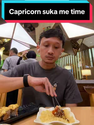 A man seated at a table in a restaurant setting, focused on his food with chop sticks. A text overlay reads "Capricorn suka me time". The background shows other people, partially obscured, and the interior of the restaurant, lit by overhead and table lamps.