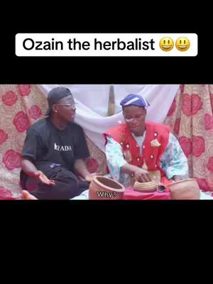 The image features two individuals in a domestic setting, likely a room with patterned fabrics. One is wearing a black t-shirt and cap while facing left, the other is wearing traditional attire and has a head wrap, and they are interacting with each other near earthenware.  An overlay at the top reads 'Ozain the herbalist' along with smiling emojis.