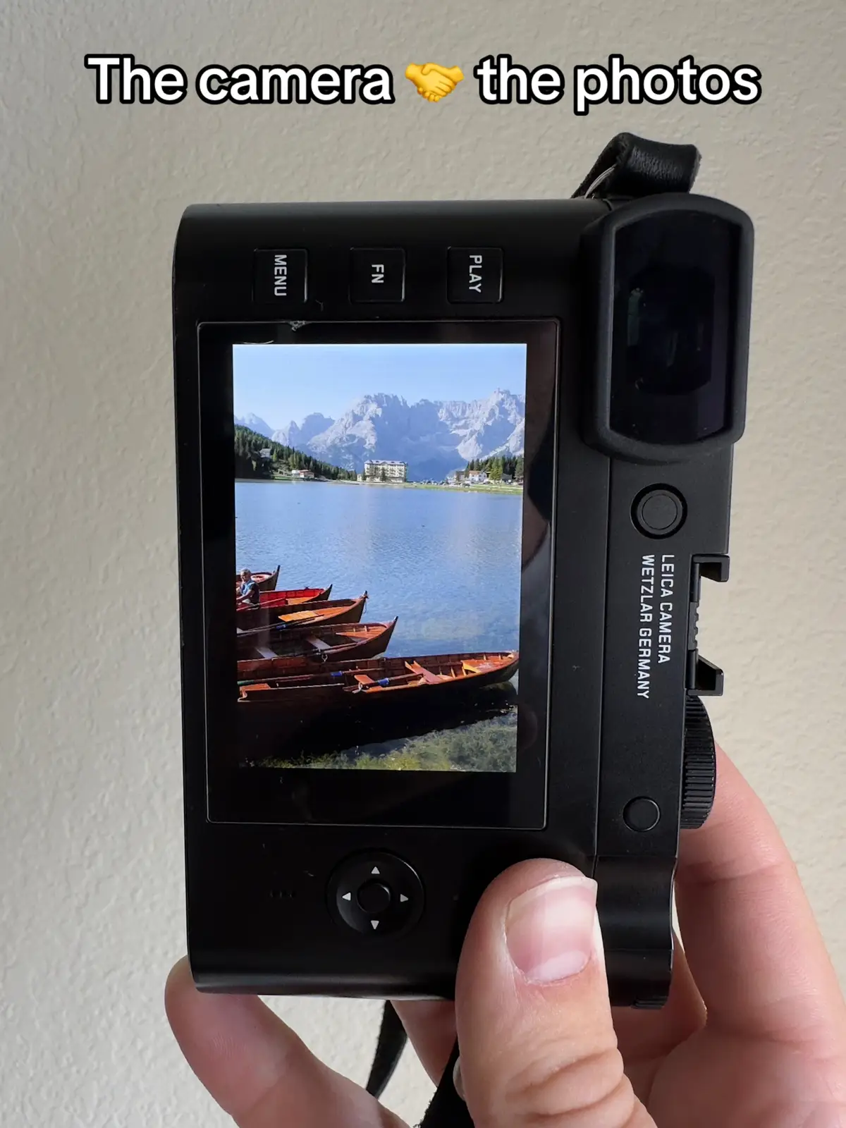 A person holding a black Leica Q2 camera, presenting its screen. The screen displays a vibrant landscape scene with mountains, a lake, and several small boats in the water, set against a bright blue sky. The handle and buttons are clearly visible, and the camera is held in a neutral, indoor setting.