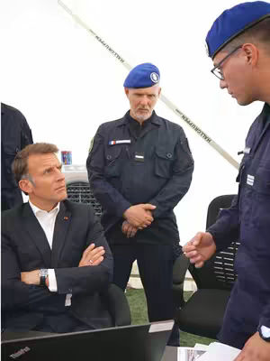 A photograph shows a group of men involved in what appears to be a discussion on the field. The main focus is on a man, likely a political figure or government official, who is seated and looking intently at something, possibly a laptop. Another man, possibly a military or security official, stands behind him with a serious expression. A third man, wearing a blue beret, looks on as well. The lighting suggests an outdoor or temporary structure, perhaps a tent. The overall tone is serious, indicating a discussion of public affairs or a sensitive situation.