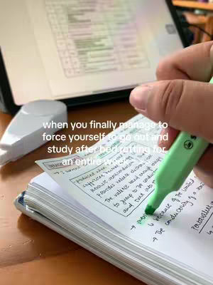 A hand with a green highlighter highlighting notes on a notepad. In the background is a tablet showing a list of items. The background also contains a white object, and a wooden desk.