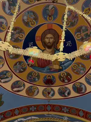 The image shows the inside of a church with a detailed ceiling. At the center, there is a circular painting of Jesus with a golden halo. Around Jesus, there are smaller circular portraits of saints and religious figures. Decorative gold elements and a chandelier-like structure are visible, along with intricate architectural patterns in the ceiling.