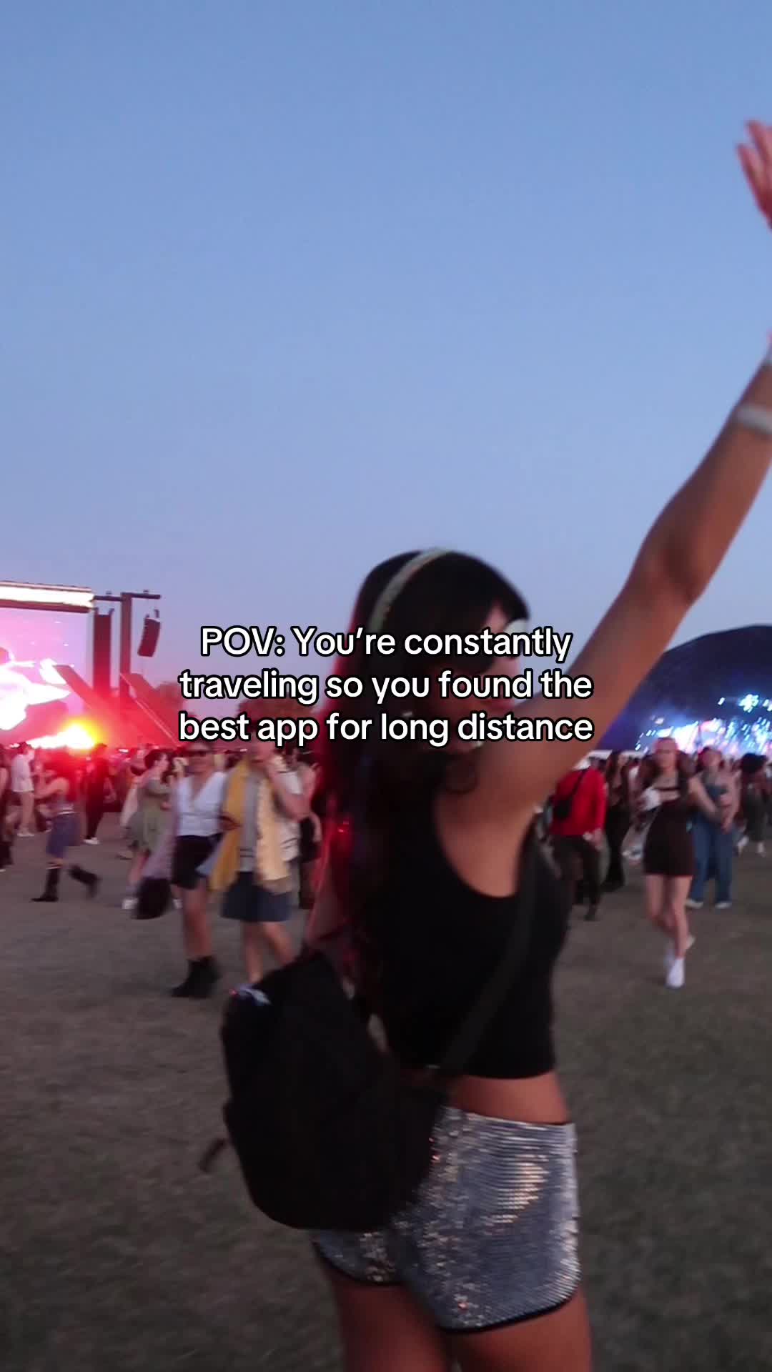 The image shows a woman with long dark hair, wearing a black tank top and sparkly shorts, walking in a crowd, seemingly at a festival. She has her right arm raised in the air, and the background is a blurred view of other people and a stage with bright lights.
