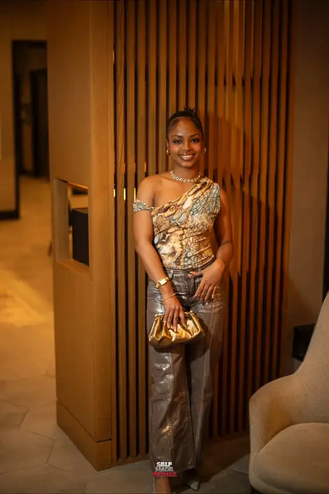 A woman in a stylish outfit poses in an upscale setting. She wears an asymmetrical top with a snake-skin-like pattern, metallic pants, and carries a gold clutch. The background includes a wooden slatted wall, and warm lighting.