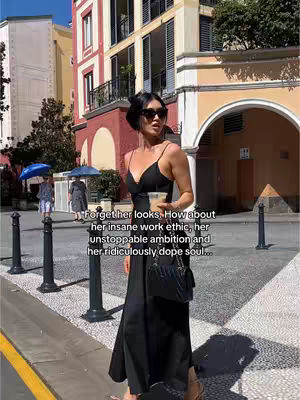 A woman in a black dress and sunglasses stands on a city sidewalk, holding a beverage. The background features European-style buildings and a sunny atmosphere.
