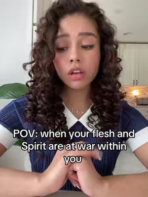 A young woman with curly brown hair looks directly at the camera with a surprised expression. She is wearing a navy blue collared shirt. The background appears to be a well-lit room with white cabinets and neutral tones.