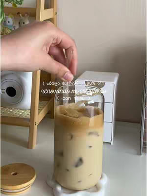 The image is a medium shot featuring a hand holding a straw over a tall glass of iced coffee. The coffee has a layered appearance, with a darker, foamy top and a lighter, milky base containing visible ice cubes. The glass sits on a white, circular coaster. Behind the glass, a white, small chest of drawers and a wooden shelf with decorative items can be seen. The background has a light, neutral color palette.