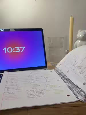 The image shows a workspace setup for studying. A laptop is open, displaying a purple and red gradient screen with the time '10:37'. In front of the laptop is a notebook with handwritten notes. Beside, a glass of water and a candle, with a small decorative frog figurine resting on the notebook.