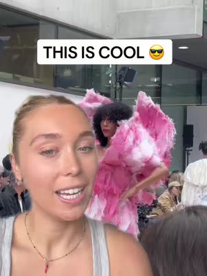 A woman with blonde hair is smiling at the camera, with a background featuring people, a fashion show attendee wearing a pink dress, and a text overlay. The woman is wearing a gray top and a necklace, with a focus on her face. The background is blurred, suggesting activity and depth.