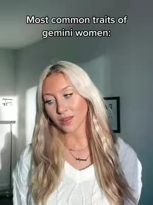 A woman with long blonde hair stands in a well-lit room. Text overlays at the top say, "Most common traits of Gemini women." The background is simple, with a framed picture visible.  The woman is wearing a white sweater and a necklace.