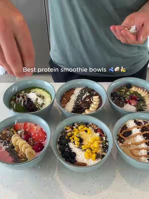 A person is holding a plate of six smoothie bowls with toppings. The person is out of focus with the bowls being the focus in the image. The bowls are arranged on a white surface, likely a countertop. The bowls have a variety of colors, toppings, and ingredients, creating visual appeal.