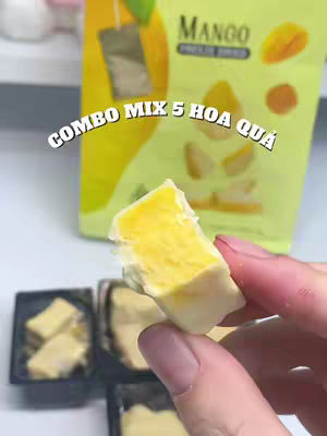 A hand holding a piece of mango candy or a similar product, with a blurred background showing a package of the product. The text overlay reads 'COMBO MIX 5 HOA QUA'.