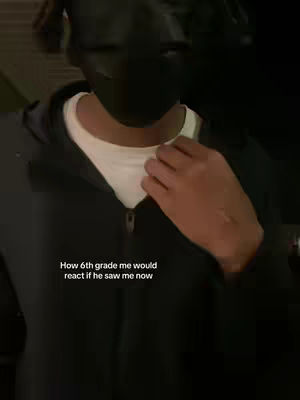 A person wearing a black mask and a black hoodie, with a white shirt visible underneath. The person's right hand is touching their chest. Text overlayed states, 'How 6th grade me would react if he saw me now'.