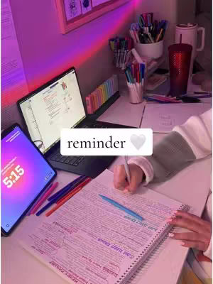A desk workspace with a laptop, tablet, and open notebook. The desk is bathed in pink and purple light, with various stationery items scattered around.