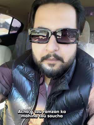 A man wearing sunglasses is positioned in the front seat of a car. He has a well-groomed mustache and beard. He is also wearing a heavy black vest over a maroon shirt with a collar. The background faintly shows the interior of a car.