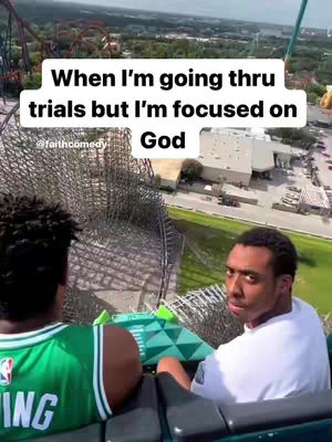 Two men are on a rollercoaster, viewed from their perspective. One, in a green Celtics jersey, faces away. The other looks directly at the camera with a concerned expression. A white text overlay states, 'When I'm going thru trials but I'm focused on God.' The background shows a cityscape with trees and buildings.