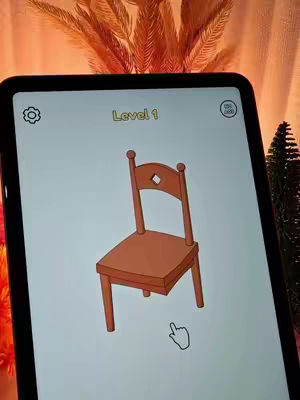 A mobile device displaying an interactive puzzle game. The game screen dominates the image, showing a drawing of a chair in an incomplete state, with a missing element. A hand cursor hovers near the bottom. The device is set against a soft, orange-toned background, likely a domestic setting, with the overall composition being clean and game-focused.