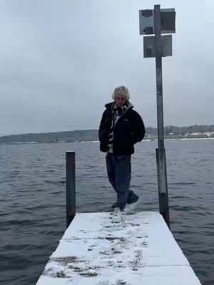 A person stands on a snow-covered pier, with a backdrop of a lake and overcast sky. The person is wearing a black jacket, jeans, and a scarf. A metal sign post is visible on the right side.