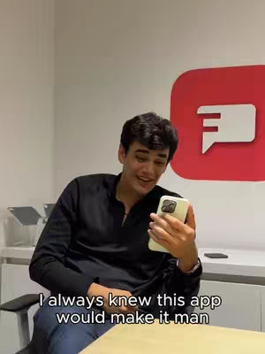 A young man with short, dark hair is seated indoors, holding a smartphone in his right hand. He's wearing a dark, zip-up shirt. His expression is a smile. The background shows a white wall with a large red logo with the app’s logo.