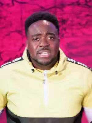A medium shot shows a dark-skinned man against a vibrant pink backdrop. He wears a yellow and black zip-up jacket with white stripes along the shoulders. The man has short, dark hair and a slight beard. His expression appears serious as if he's speaking or about to speak. The background is a solid pink, with a subtly textured pattern creating some visual interest.