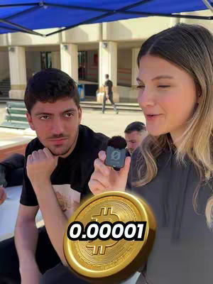 A woman and a man are standing outdoors with a Bitcoin-like coin overlaid on the bottom of the image to show a numerical value of 0.000001. The woman is holding a microphone; the man is looking to the woman. Other people are in the background of an outdoor scene, under a blue tent structure.