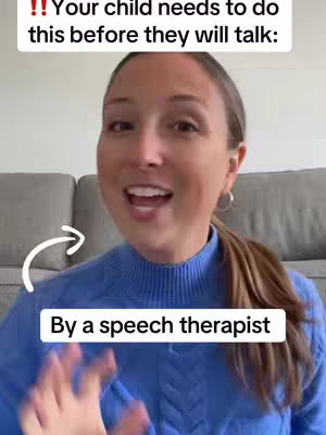 A woman is centered in the frame, smiling and speaking. She wears a medium blue sweater. In the background is a light gray couch. Text is added to the image.