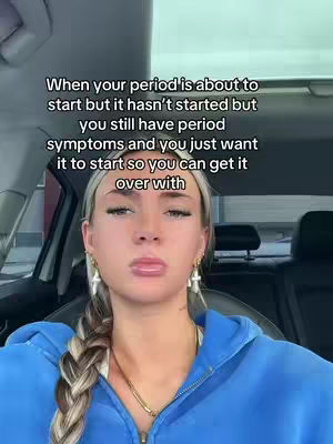 A selfie-style shot taken from inside a car. A fair-skinned woman with blonde hair styled in a braid and wearing noticeable earrings and a gold necklace is in the foreground. She has a subtle expression, with slightly pursed lips. She is wearing a blue hoodie. The background is blurred, but visible through the windshield are elements of the car's interior and outdoor scenery.