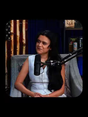 The cover image features a woman in a seated position, centered and against a dark background. The woman is the primary focus, dressed in a white sleeveless top. She has dark hair and a light skin tone. She is seated on a grey armchair, with a microphone and a stand to her right. Behind her, there's an American flag and a dark blue panel. The image has a cinematic feel due to the lighting and arrangement.