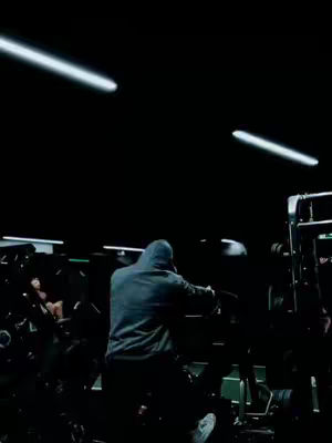 A person in a dark gym setting wearing a hoodie is working out on a machine. The background is dark with some equipment visible and two bright linear lights on the ceiling.