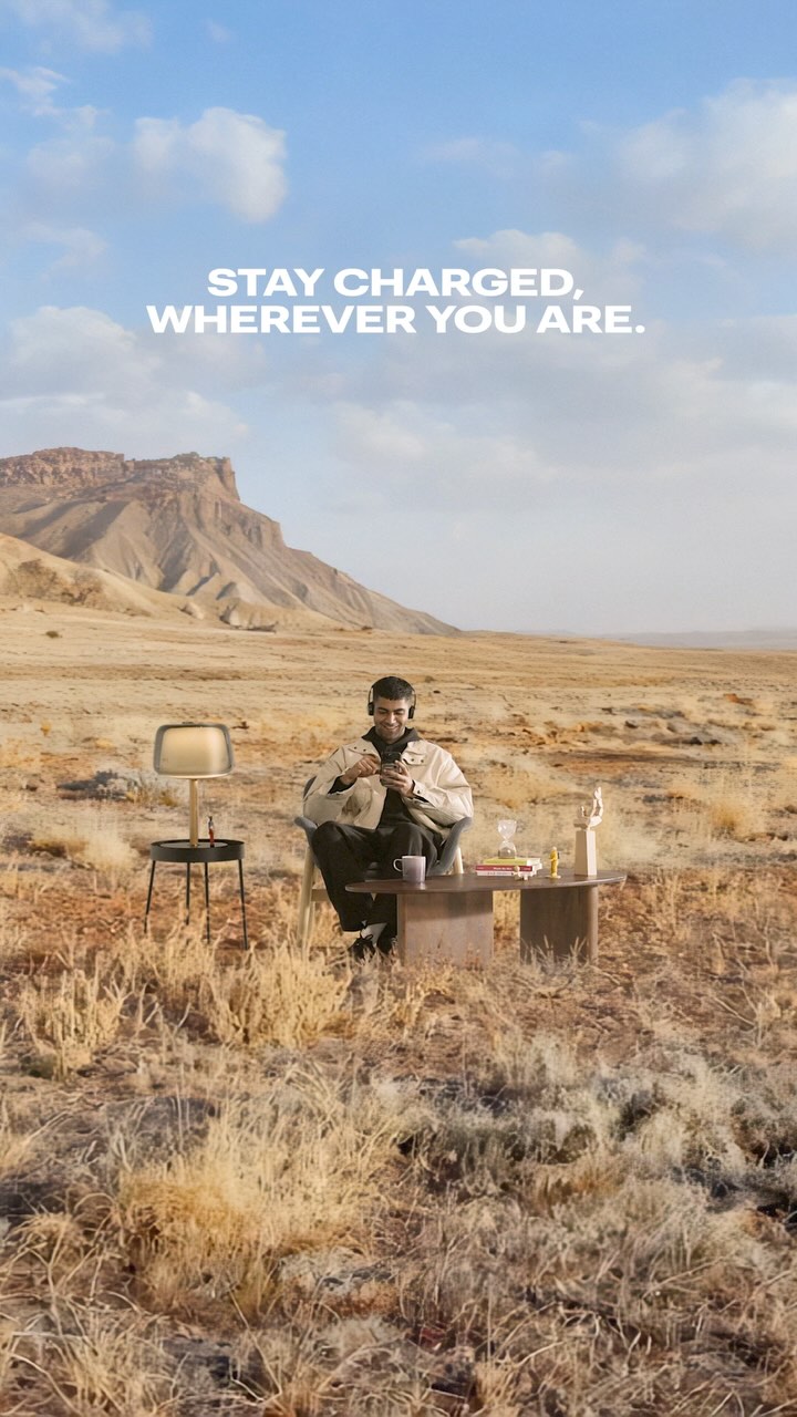 A man sits at a desk with a lamp, books, and a sculpture in a desert landscape under a blue sky. White text overlays in the top, reading "STAY CHARGED, WHEREVER YOU ARE."
