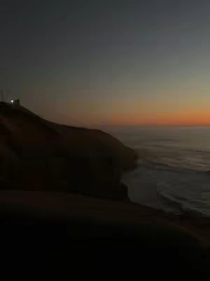 An image of a coastal scene at dusk. The foreground is a dark, silhouetted cliff or hillside, with rough texture and subtle variations in tone. Above it, the sky transitions from a deep, dark blue at the top to a warm orange and yellow towards the horizon. The sea is visible to the right, with gentle waves and a light reflecting the colors of the sky. A structure with a few lights stands on the cliff in the far distance.