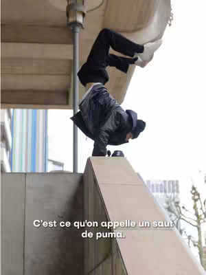 A person doing a handstand on a concrete structure. The person is wearing black clothing and white shoes. A text overlay at the bottom says, “C’est ce qu’on appelle un saut de puma.”