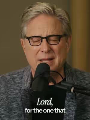 A mid-shot of a man wearing glasses and a dark gray jacket, recording himself with a microphone. Text overlayed reads, "Lord, for the one that." The background is blurred.