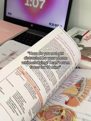 The image shows a close-up of an open textbook filled with anatomical diagrams and notes, suggesting a study environment. The book is angled towards the viewer, with a laptop in the background displaying a digital clock. Additional books and papers are visible, creating a busy desk scene.