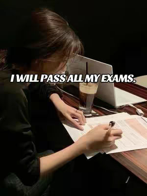 A person is sitting at a wooden table, writing on a piece of paper with a pen. A laptop and a glass with an iced drink and straw are on the table. The text overlay says, "I WILL PASS ALL MY EXAMS."