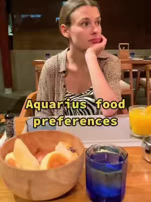 A woman with light skin sits at a table in a restaurant setting with one arm resting on the table and her chin resting on her hand. There is an array of food and drink in the foreground including a wooden bowl filled with sliced fruit and a glass of blue liquid.